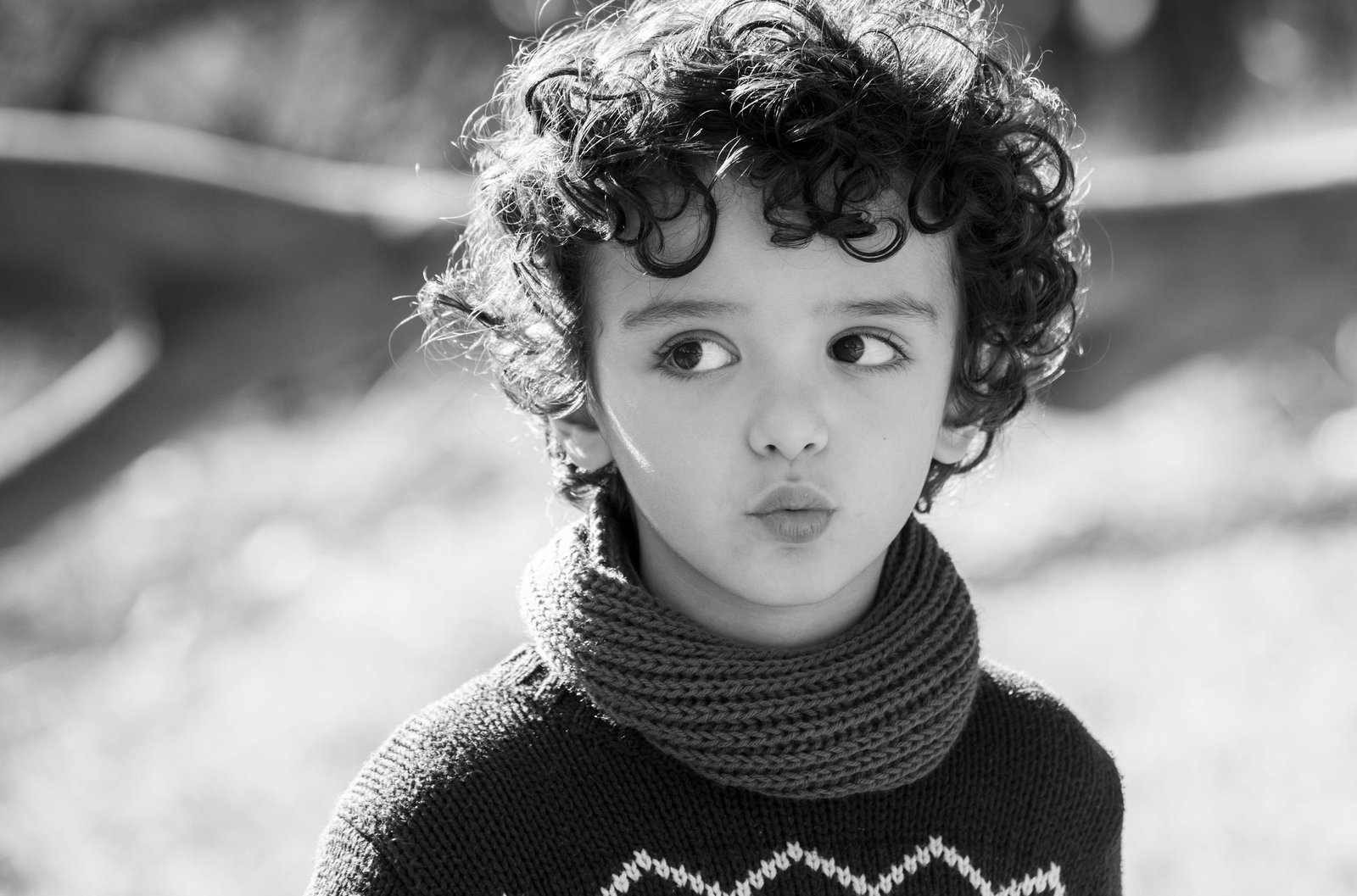 Monochrome image of a child with curly hair wearing a scarf and sweater outdoors.