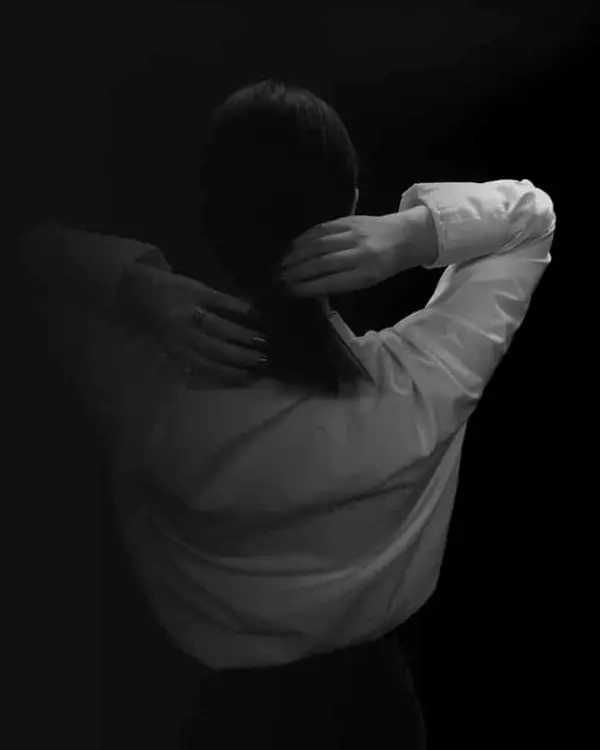 Femme de dos en chemise blanche, les mains dans les cheveux, sur fond noir