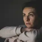 Femme au regard pensif, les bras croisés sous le menton, dans une pièce sombre