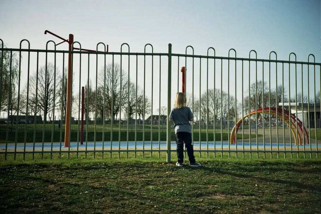 Jeune enfant seul qui regarde une aire de jeux vide à travers une grille