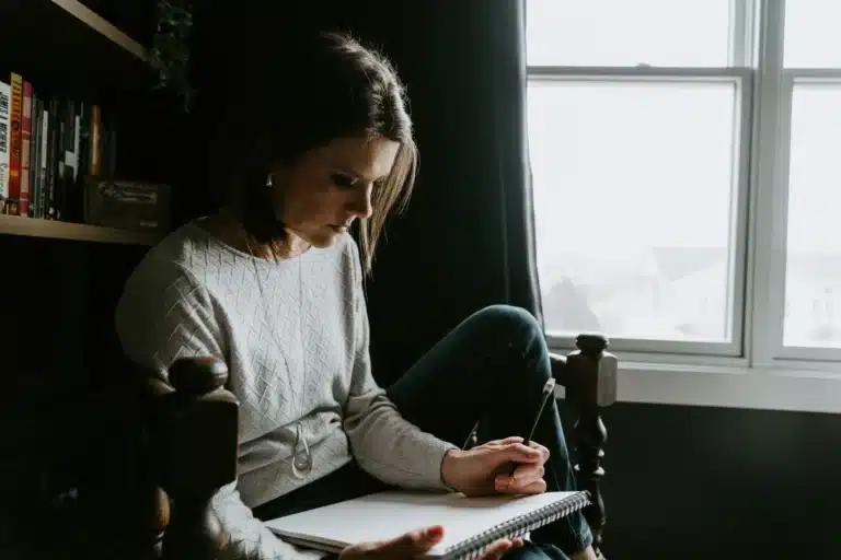 Femme qui écrit dans un carnet, assise dans un fauteuil près de la fenêtre
