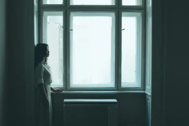 Femme seule debout devant une grande fenêtre dans une pièce sombre