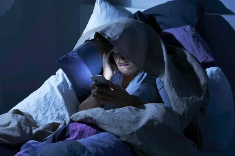 Femme blottie sous sa couette la nuit, éclairée par son téléphone