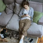 Jeune femme allongée sur son canapé, absorbée par son téléphone dans un salon en désordre