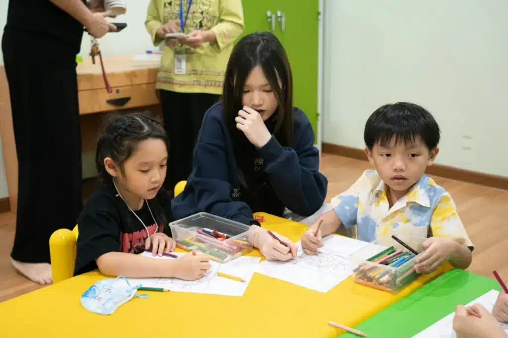 Des enfants dessinent autour d’une table avec leur enseignant, dans une classe lumineuse